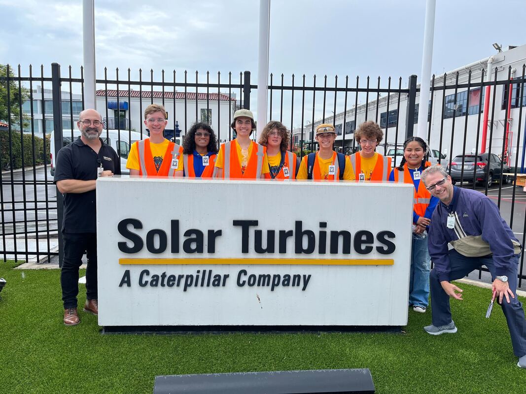 7 high school students wearing orange safety vests pose standing behind an exterior Solar Turbines sign with two employees